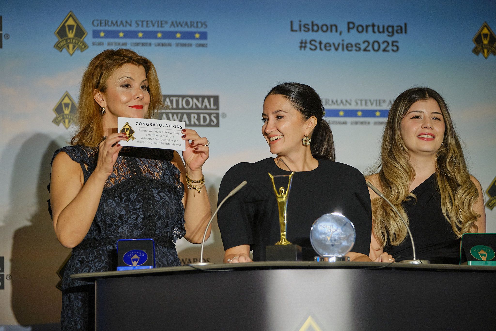 Tres ganadoras en el atril, una sosteniendo una tarjeta de felicitación, en los International Business Awards® 2025