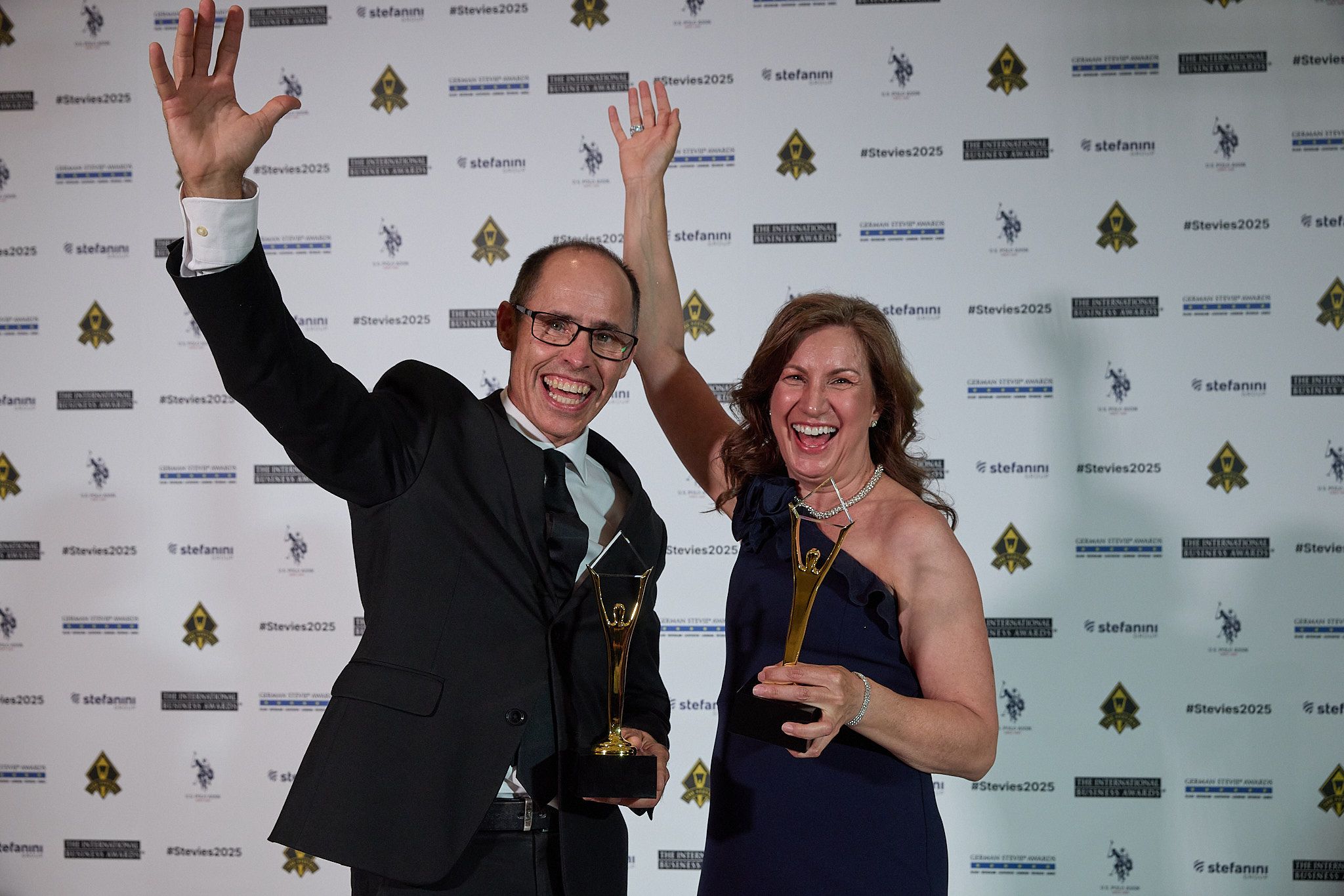 Dos ganadores celebrando con los brazos en alto y sus trofeos Stevie de oro en los International Business Awards® 2025