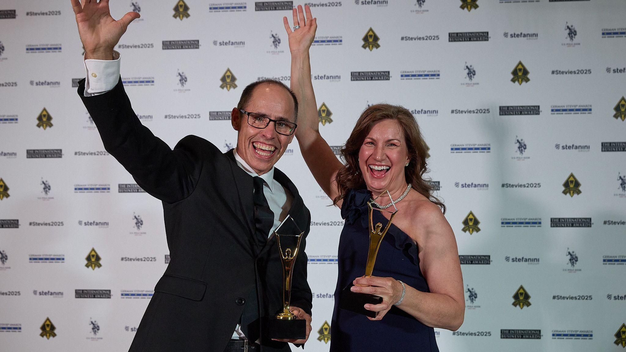 Dos ganadores celebrando con los brazos en alto y sus trofeos Stevie de oro en los International Business Awards® 2025