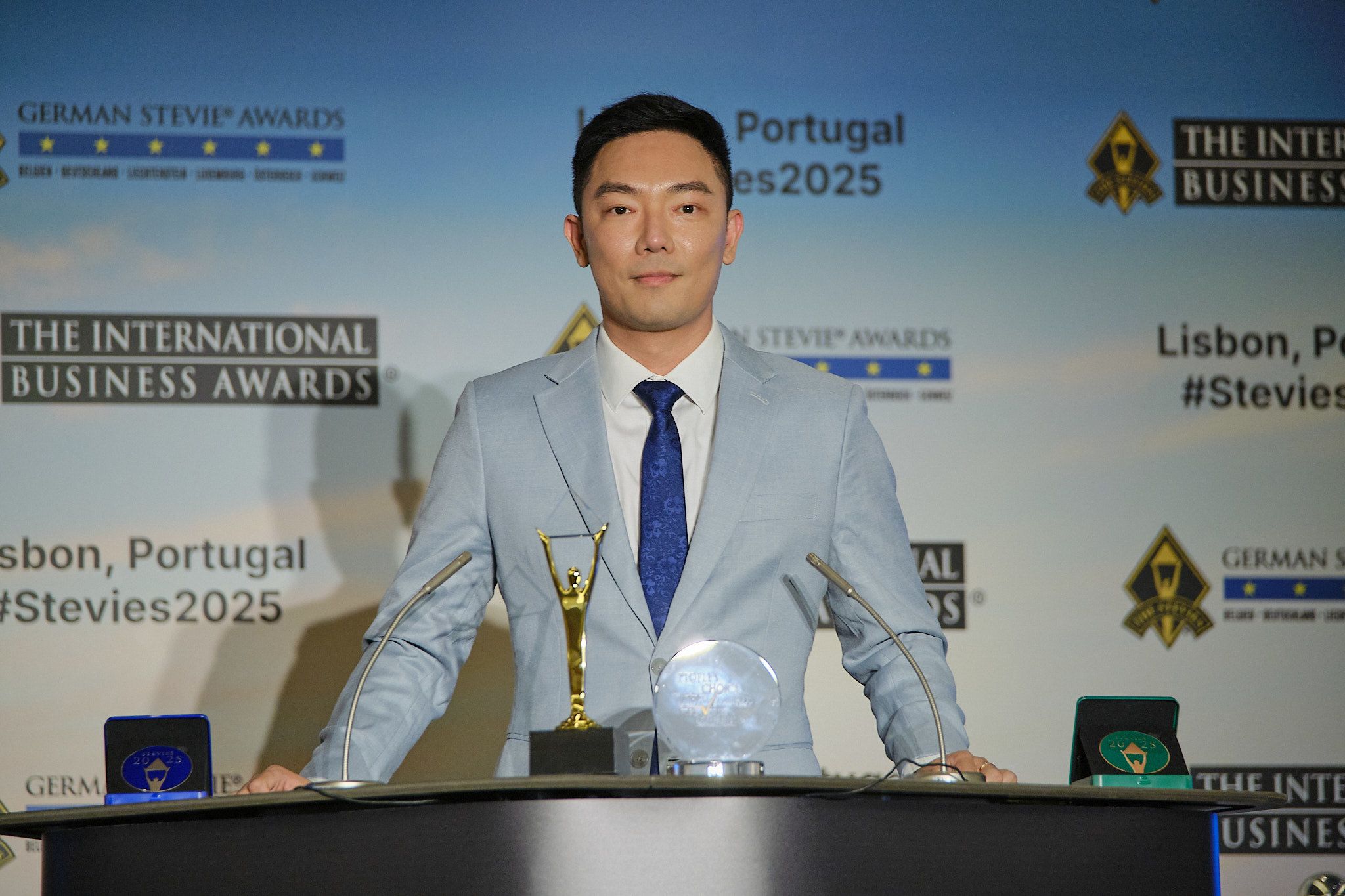 Ganador con traje azul claro y su trofeo Stevie de oro en el atril de los International Business Awards® 2025