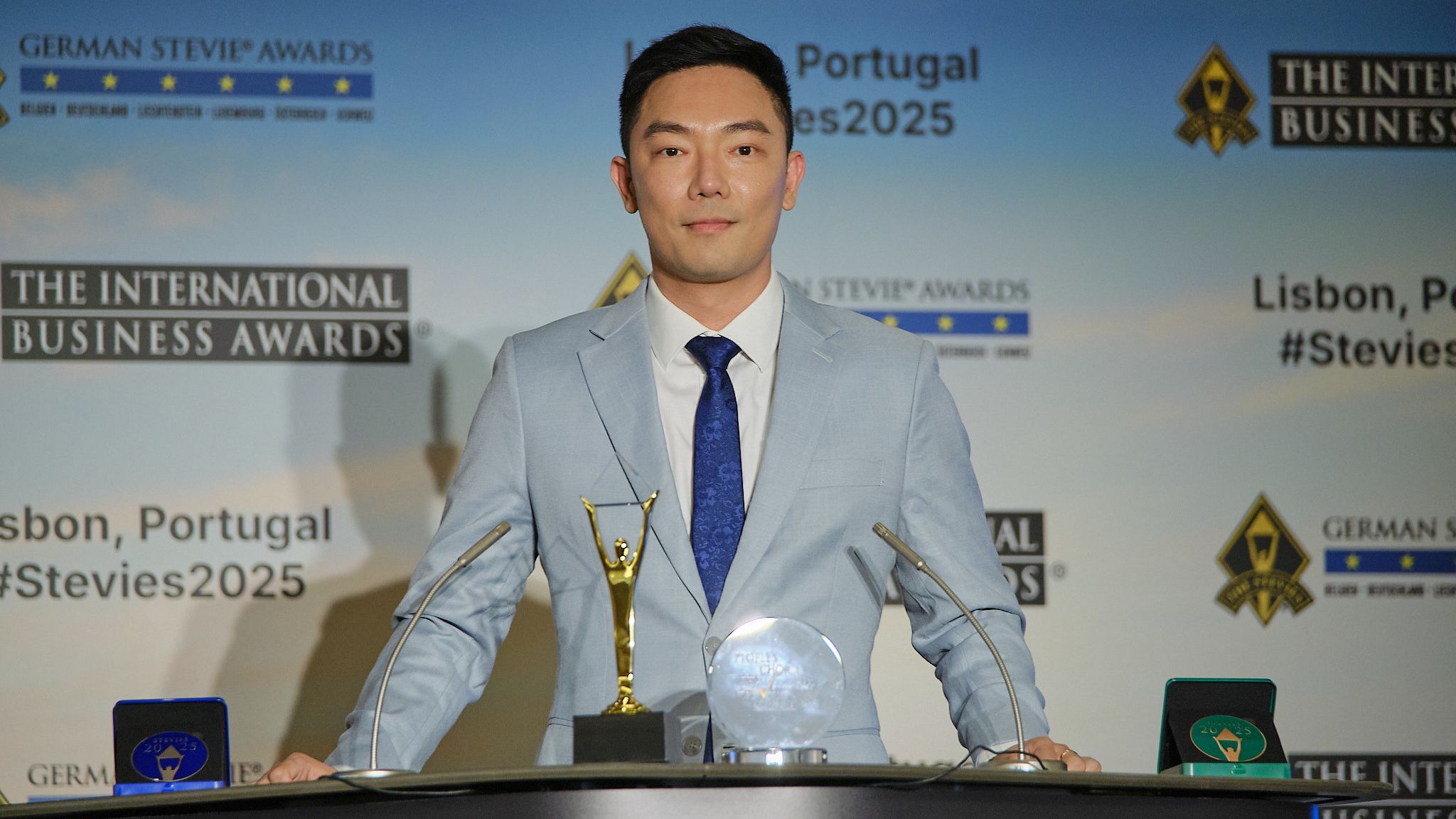 Ganador con traje azul claro y su trofeo Stevie de oro en el atril de los International Business Awards® 2025