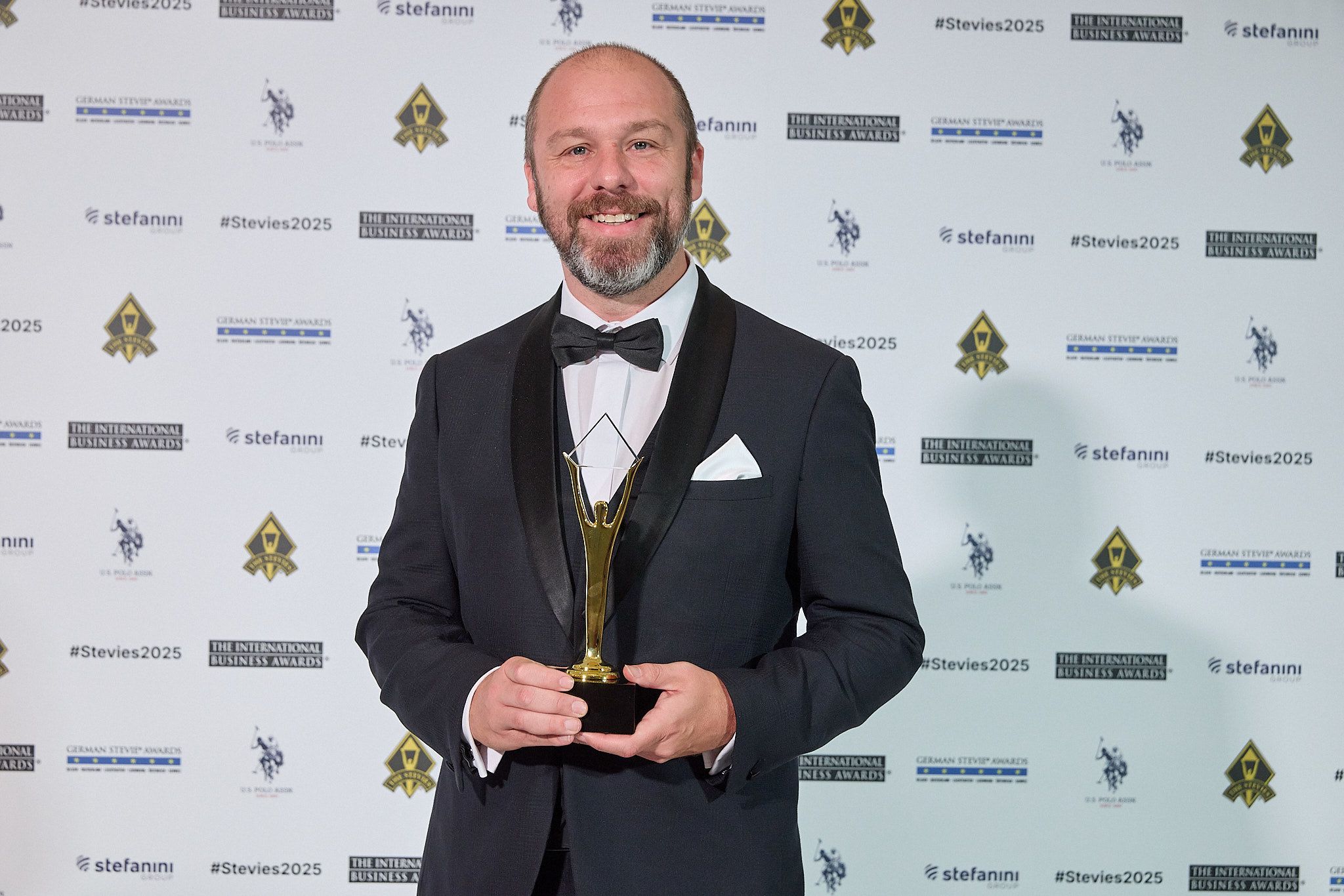 Ganador en esmoquin sonriendo con su trofeo Stevie de oro en la alfombra roja de los International Business Awards® 2025