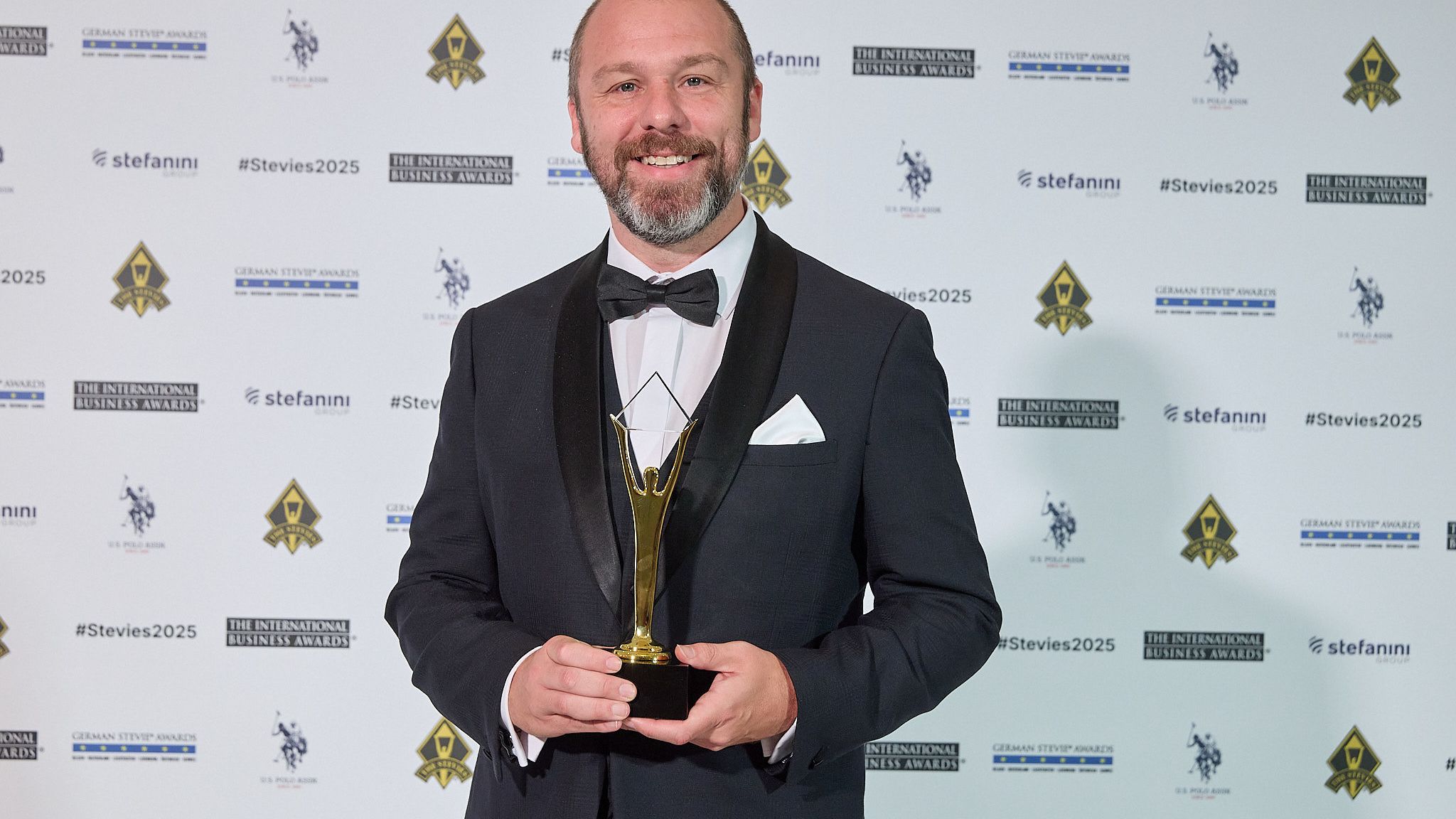 Ganador en esmoquin sonriendo con su trofeo Stevie de oro en la alfombra roja de los International Business Awards® 2025